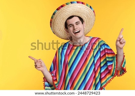 Young man in Mexican sombrero hat and poncho pointing at something on yellow background