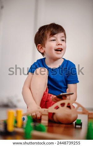 A joyful young boy with a smartphone sits amidst a wooden train setup, his bright laughter echoing the playful fusion of digital and tangible play Royalty-Free Stock Photo #2448739255
