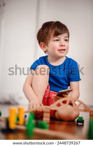 A joyful young boy with a smartphone sits amidst a wooden train setup, his bright laughter echoing the playful fusion of digital and tangible play Royalty-Free Stock Photo #2448739267