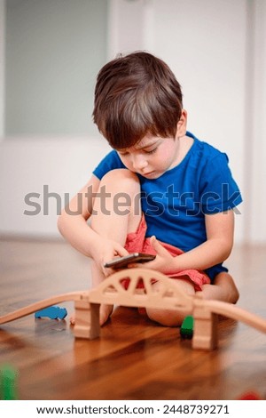 A joyful young boy with a smartphone sits amidst a wooden train setup, his bright laughter echoing the playful fusion of digital and tangible play Royalty-Free Stock Photo #2448739271