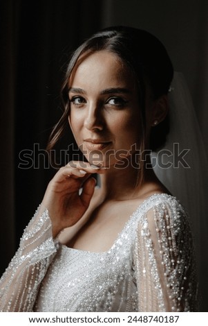 Stunning bride with dark hair and bright smile in a white lace wedding gown and long veil, standing elegantly by a window on her special day. The low-angle shot captures her grace and beauty.