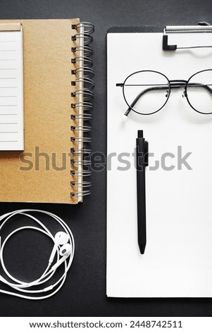 Blank sheet of paper next to pen, notepads, white wired headphones and glasses on black background