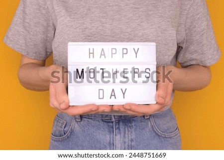 Happy Mothers Day. Female hands hold lightbox with letters in front of yellow background. 