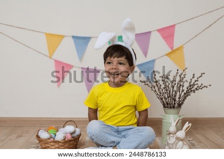 A child sits next to a basket and colorful eggs for Easter.Easter photo.Child wearing Easter bunny bunny ears laughs