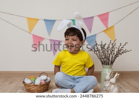 A child sits next to a basket and colorful eggs for Easter.Easter photo.Child wearing Easter bunny bunny ears laughs