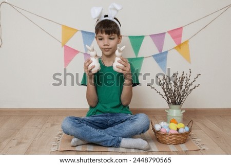 Easter photo shoot.A child wearing bunny ears holds an Easter bunny in his hands.Multi-colored Easter eggs lie in abasket and child sits next to him.