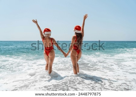 Women Santa hats ocean play. Seaside, beach daytime, enjoying beach fun. Two women in red swimsuits and Santa hats are enjoying themselves in the ocean waves and raising their hands up.