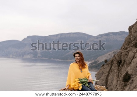 Portrait happy woman woman with long hair against a background of mountains and sea. Holding a bouquet of yellow tulips in her hands, wearing a yellow sweater