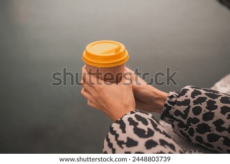 A person holding a coffee cup with a yellow lid. The person is wearing a leopard print jacket.