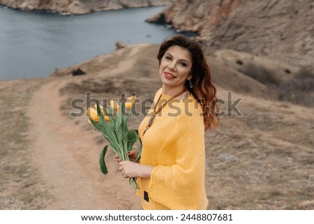 Portrait of a happy woman with long hair against a background of mountains and sea. Holding a bouquet of yellow tulips in her hands, wearing a yellow sweater