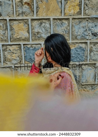 A pakistani simple girl posing for a picture while wearing traditional dress and jewellery , having henna on her hands with a beautiful background of stone wall .