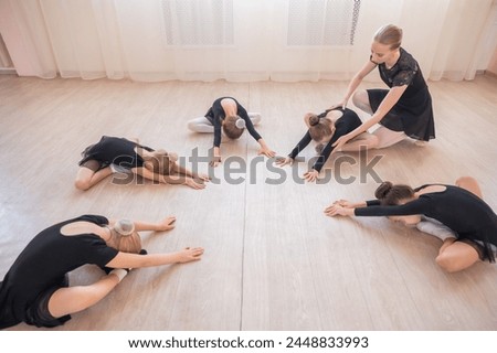 Caucasian woman and five little girls sit in a circle and do stretching at a ballet school. 
