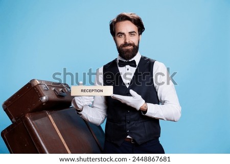 Hotel employee leading tourists in the right way using reception wall sign. Skilled professional bellboy posing on camera and holding front desk indicator, guiding guests and travellers.