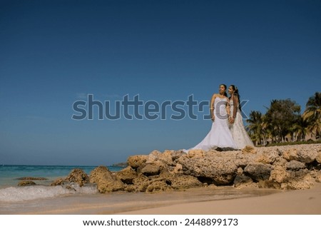 newly married women wearing their wedding dresses hugging on a giant rock on the shore of the beach 