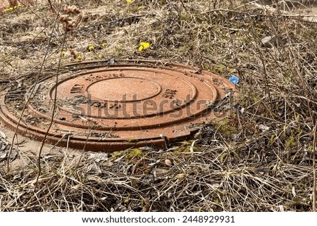 A rusty iron hatch with a pattern covered dirt above an observation well is located next to a dry grass in the daytime. Close-up