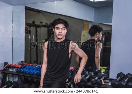 A young, skinny, and lanky Asian man, dressed in a black tank top, is in the gym for his first training session, showing determination. Royalty-Free Stock Photo #2448986141