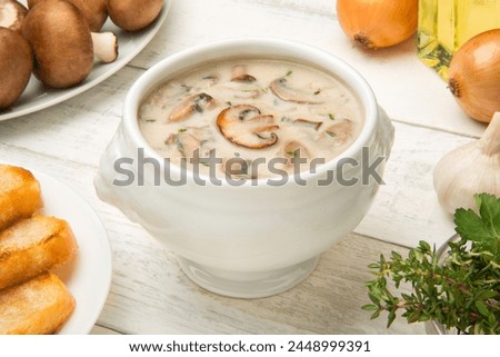 Cream of mushroom soup in a white porcelain tureen on a white rustic table with ingredients. Selected focus. Royalty-Free Stock Photo #2448999391