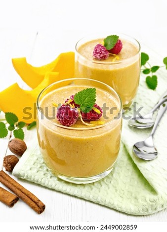Yogurt, pumpkin and raspberry dessert with nutmeg, cinnamon, orange zest in two glasses, a vegetable, spoons and spices on a napkin against white wooden board background