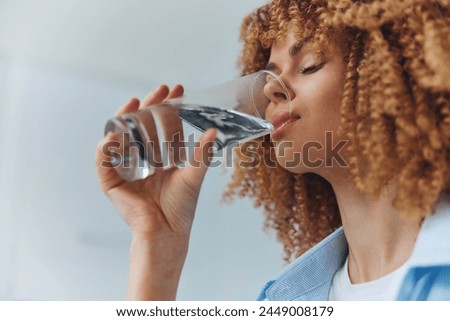 Woman with Curly Hair Enjoying a Refreshing Drink of Water in a Glass in a Brightly Lit Room Royalty-Free Stock Photo #2449008179