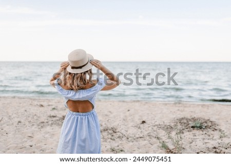 Portrait of stylish female on sand. Beautiful girl standing on beach ocean and enjoying sunny summer happy day on vacation. Back view. Young attractive blond woman in straw hat joyfully walks near sea Royalty-Free Stock Photo #2449075451