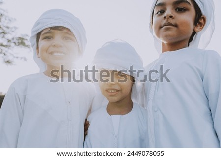 Children playing together in Dubai in the park. Three children with dish dasha look into the camera smiling. Emirati people wearing traditional kandura white dress. Royalty-Free Stock Photo #2449078055