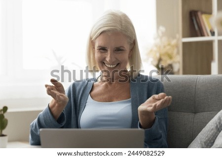 Overjoyed older female share news during on-line talk to family, resting on sofa looking at laptop screen, gesturing, lead pleasant conversation remotely using computer and videoconference application Royalty-Free Stock Photo #2449082529
