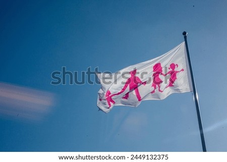 Flag where picture of parents with children. Pink sign on white flag. Blue sky background.