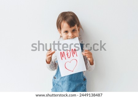 little cute white brunette girl 1.5 years old holds in her hands and teeth a card with the inscription text mom and a red heart on a white background, the concept of congratulations on mother's day.
