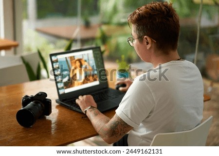 Professional photo editor works on laptop in bright coworking space. Tattooed, stylish person edits image, focused on screen, camera beside. Modern freelance job in creative industry, design workflow.
