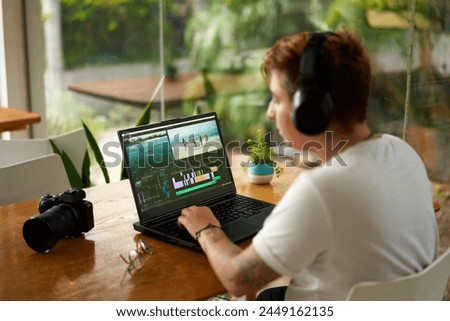 Professional videographer in headphones edits footage on laptop at coworking space. Camera gear on table, creative editing process, freelance job in modern workspace. Editor works on travel video.