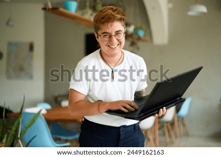 Smiling transgender professional holds laptop, works happily in modern office space. Inclusive workplace, young trans person enjoys career, experiences job fulfillment with tech in eco-friendly area.