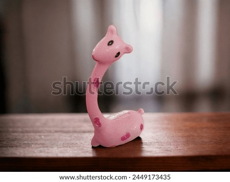 A pink plastic piggy bank with a cute giraffe with mauve dots and black eyes on a wooden table, a lighted window background, and a white curtain - a small plastic piggy bank 