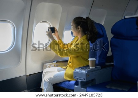 A woman is sitting on an airplane, taking a picture of the view outside the window. She is wearing a yellow jacket and white pants
