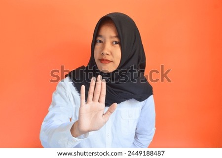 A young Asian woman in a hijab raises her hand in front of a stop gesture sign
