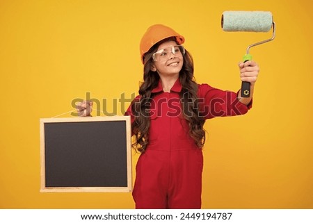 Little builder expert in helmet on construction site. Teen girl painter with painting brush tool or paint roller. Child on repairing work. Renovation concept. Happy smiling girl.
