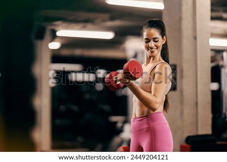 A muscular sportswoman is doing dumbbells curls in a gym and flexing muscles. Royalty-Free Stock Photo #2449204121