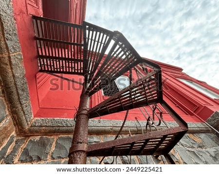 A spiral staircase is seen from the side of a building. The staircase is made of metal and is rusted. The building is red and has a stone facade. The sky is cloudy