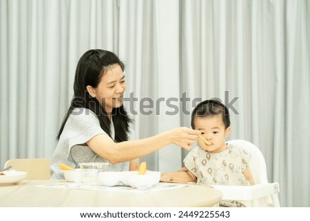 Asian mother feeding baby boy sitting on white highchair with. messy table and gray backdrop