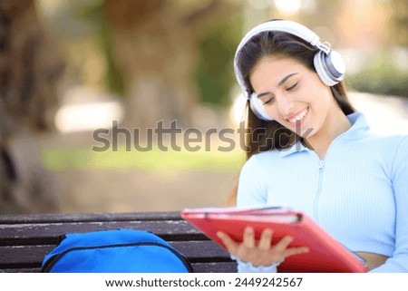 Happy student in a park listening audio guide wearing headphone and reading notes