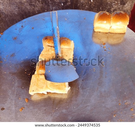 Fluffy bread bun being roasted with spices on a iron tawa-pan for preparing vada pav or wada pao with a ladle.