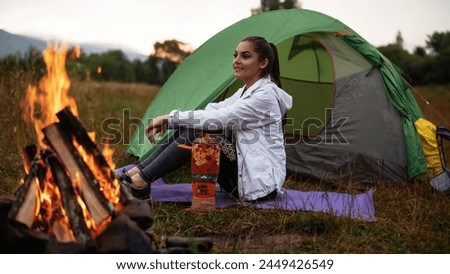 Beautiful female traveler smiling while looking at the nature and resting on grass near bonfire. Tourism concept. Stock photo 