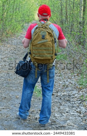 one male tourist in a red baseball cap with an olive-colored backpack on his back and dressed in a gray T-shirt walks along a road in the forest during the day