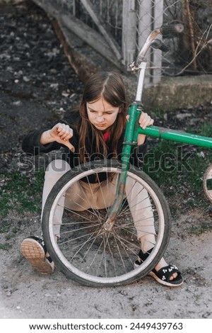 A little sad teenage girl, a dissatisfied upset child, showing a dislike, sits near an old bicycle with a broken, punctured wheel tire outdoors. Photography, portrait. Royalty-Free Stock Photo #2449439763