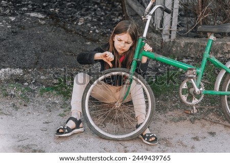 Little beautiful sad teenage girl, dissatisfied upset child showing dislike thumbs down sits near an old retro bicycle with a broken, punctured wheel tire outdoors. Photography, portrait, lifestyle. Royalty-Free Stock Photo #2449439765