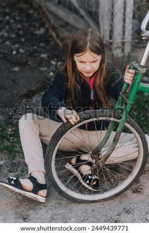 Little teenage girl, dissatisfied, distressed child sits near an old bicycle with a broken, punctured wheel tire outdoors. Royalty-Free Stock Photo #2449439771