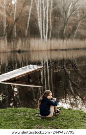 Little beautiful girl, lonely dreaming child sits on green grass against the backdrop of a lake in nature outdoors. Photography, portrait.