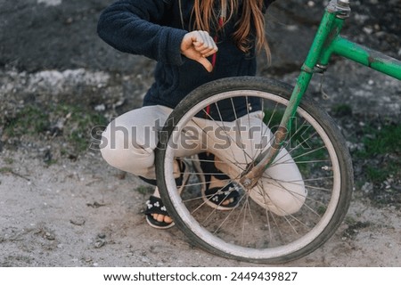 A teenage girl, a child, showing a dislike thumbs down sits near an old retro bicycle with a broken, punctured wheel tire outdoors. Photography, portrait, lifestyle. Royalty-Free Stock Photo #2449439827
