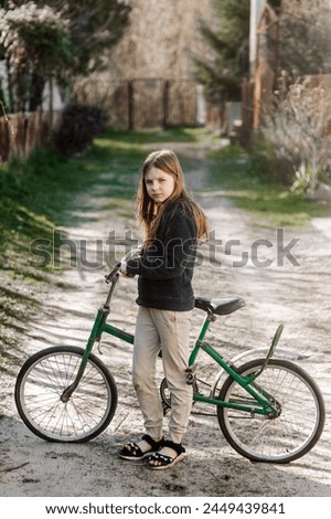 A beautiful teenage girl child stands with an old rusty bicycle in her hands on a road in the countryside, village, outdoors. Photography, portrait.