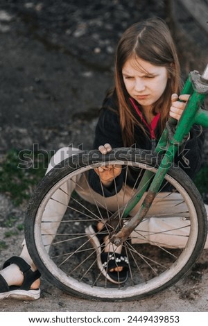 Little teenage girl, dissatisfied, distressed child sits near an old bicycle with a broken, punctured wheel tire outdoors. Royalty-Free Stock Photo #2449439853