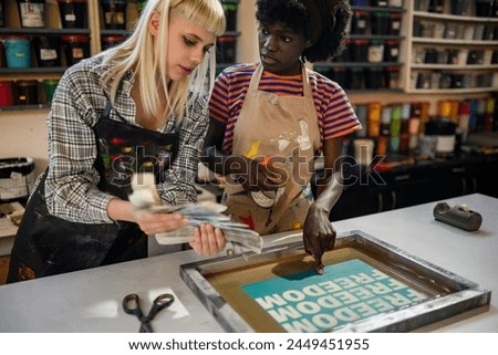 Graphic industry worker with color swatch in hands choosing colors with her multicultural colleague. African american graphic worker is pointing at silk screen printing plate and discussing process.